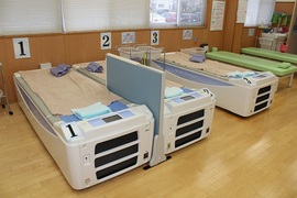 写真上） 水圧マッサージベッド『アクアラグーン』を3台導入いただいております。 （写真左） 天井のレールに掛けられたマグナーパック。 治療の効率化を図る画期的なアイデアです。